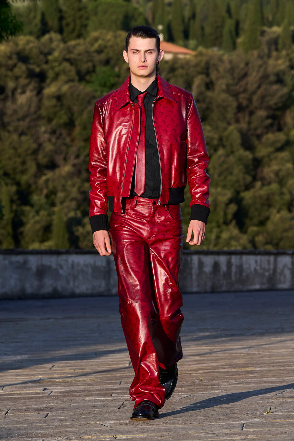 Male model in a shiny red leather suit and black shirt walks toward the camera on a rooftop terrace with greenery in the background.