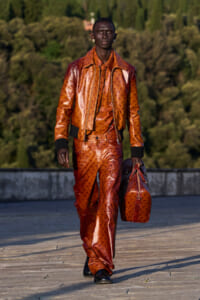 Male model walking on a rooftop runway in a matching orange-brown patterned leather suit, carrying a matching duffel bag.