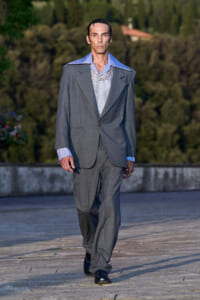 Male model in an oversized gray suit walking on a stone terrace, blue checkered shirt and chunky chain necklace visible.