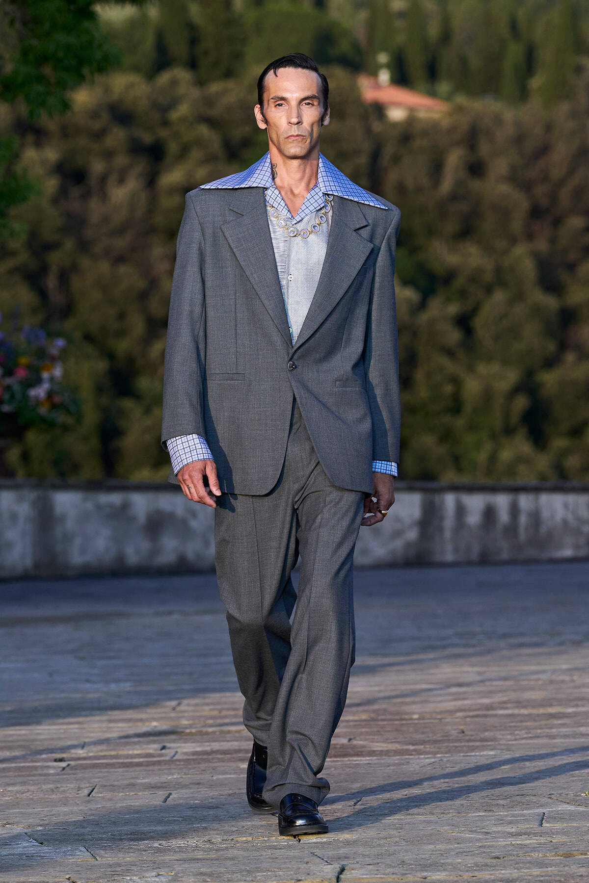 Male model in an oversized gray suit walking on a stone terrace, blue checkered shirt and chunky chain necklace visible.