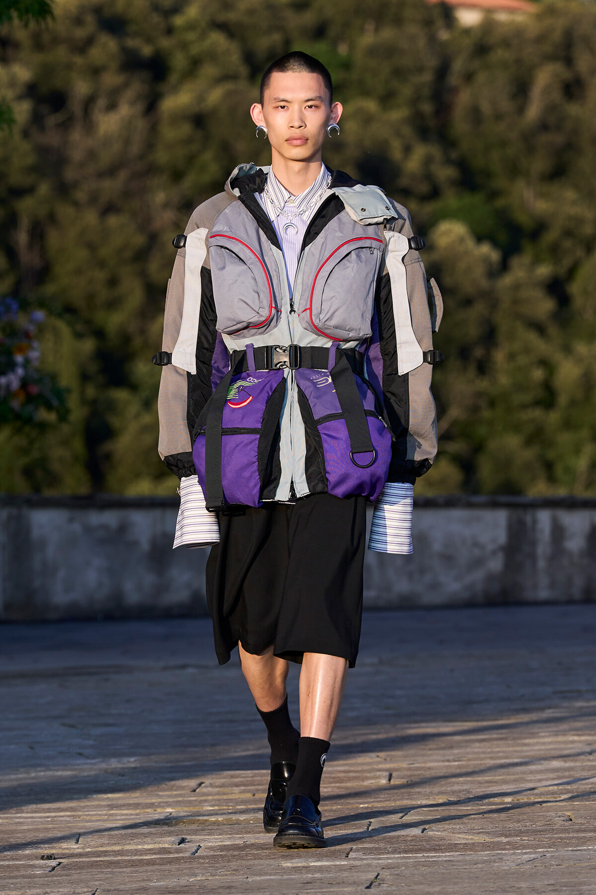 Model walking outdoors in a layered, oversized streetwear outfit with gray and purple vests and striped sleeves.