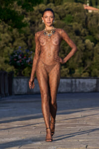 Model struts down a stone terrace in a sheer, nude-toned mesh bodysuit with gold crescent patterns and bold jewelry, outdoors.