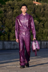 Person walking on a stone terrace wearing a shiny purple patterned jumpsuit, carrying a matching purple bag.