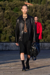 Male model walks on runway wearing a black leather jacket, sheer patterned top, black shorts, and bold jewelry, carrying a textured black duffel bag.