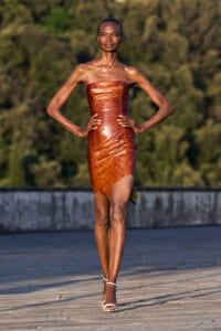 Black model walks a runway outdoors in a strapless copper-brown leather dress with a high slit and white heeled sandals, hands on hips.