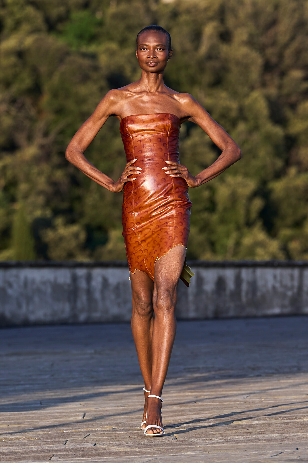 Black model walks a runway outdoors in a strapless copper-brown leather dress with a high slit and white heeled sandals, hands on hips.
