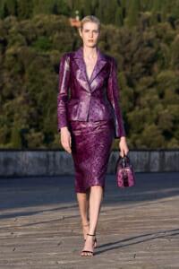 Female model wearing a shiny purple patterned suit and matching handbag, walking on a wooden deck outdoors in heels.