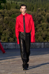 Male model walking on a runway in a bright red shirt, black tie, and black leather pants outdoors with trees in the background