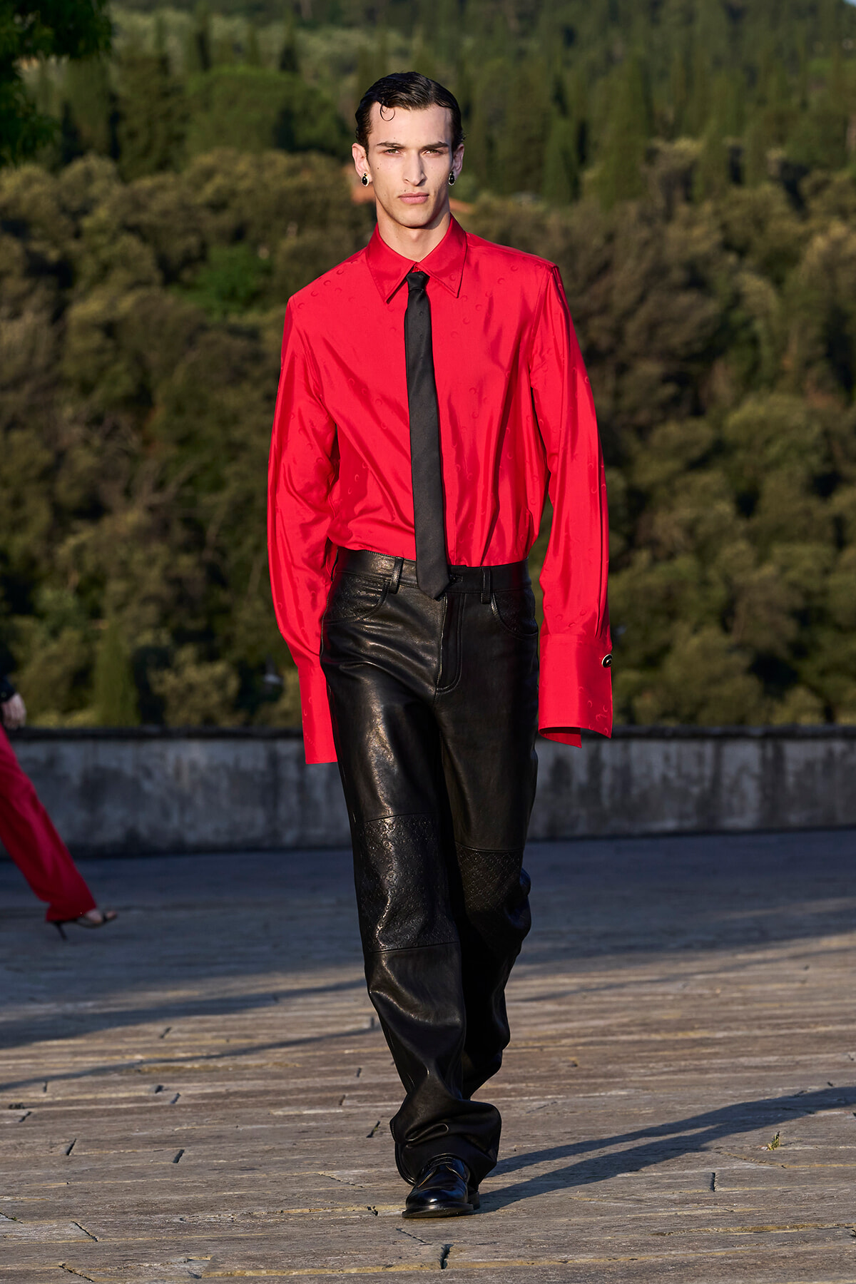 Male model walking on a runway in a bright red shirt, black tie, and black leather pants outdoors with trees in the background