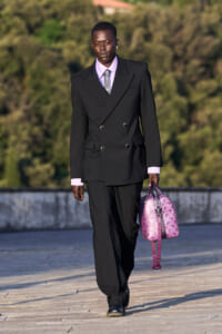 A tall model in a black double-breasted suit with lavender shirt and tie walks outdoors, carrying a lavender-purple patterned handbag.