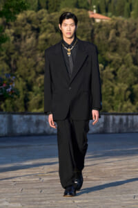 Male model walks a runway outdoors in a black suit and gold necklace against a green, tree-lined backdrop.