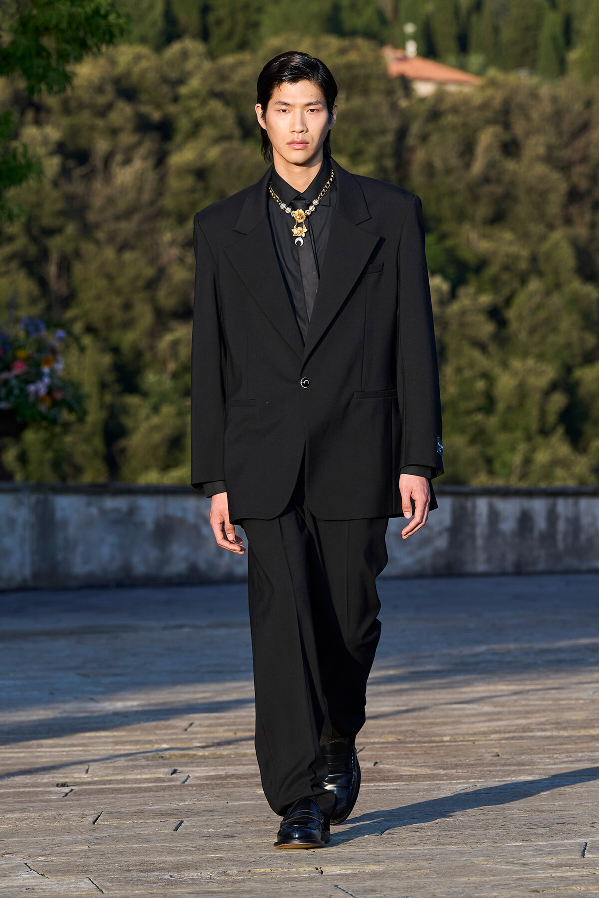 Male model walks a runway outdoors in a black suit and gold necklace against a green, tree-lined backdrop.