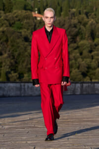 Model wearing a bright red double-breasted suit with a black shirt, walking on an outdoor runway.