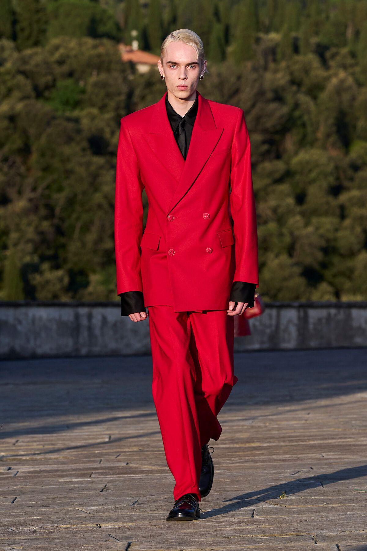 Model wearing a bright red double-breasted suit with a black shirt, walking on an outdoor runway.