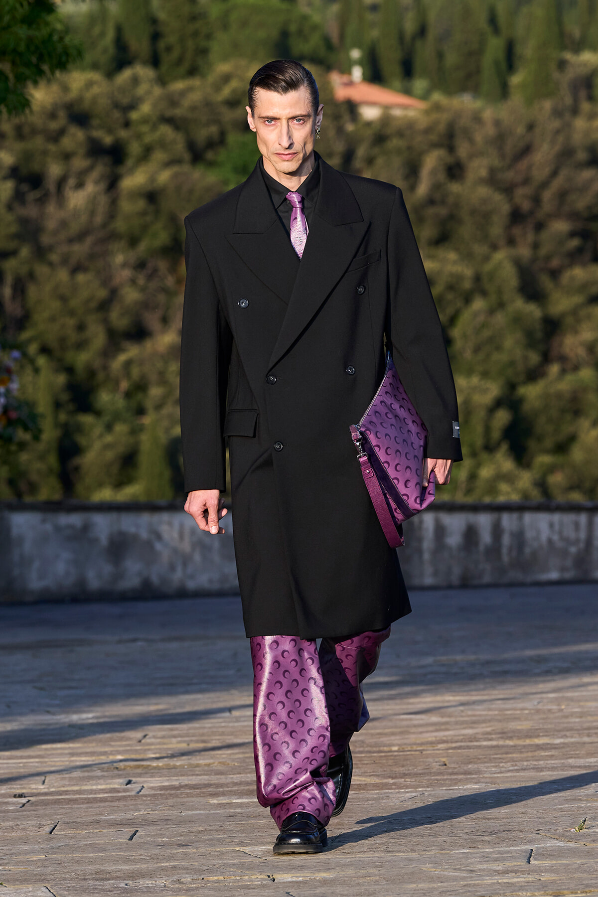 Model walks a runway in a long black double-breasted coat with purple satin-patterned pants and a matching purple clutch bag in hand