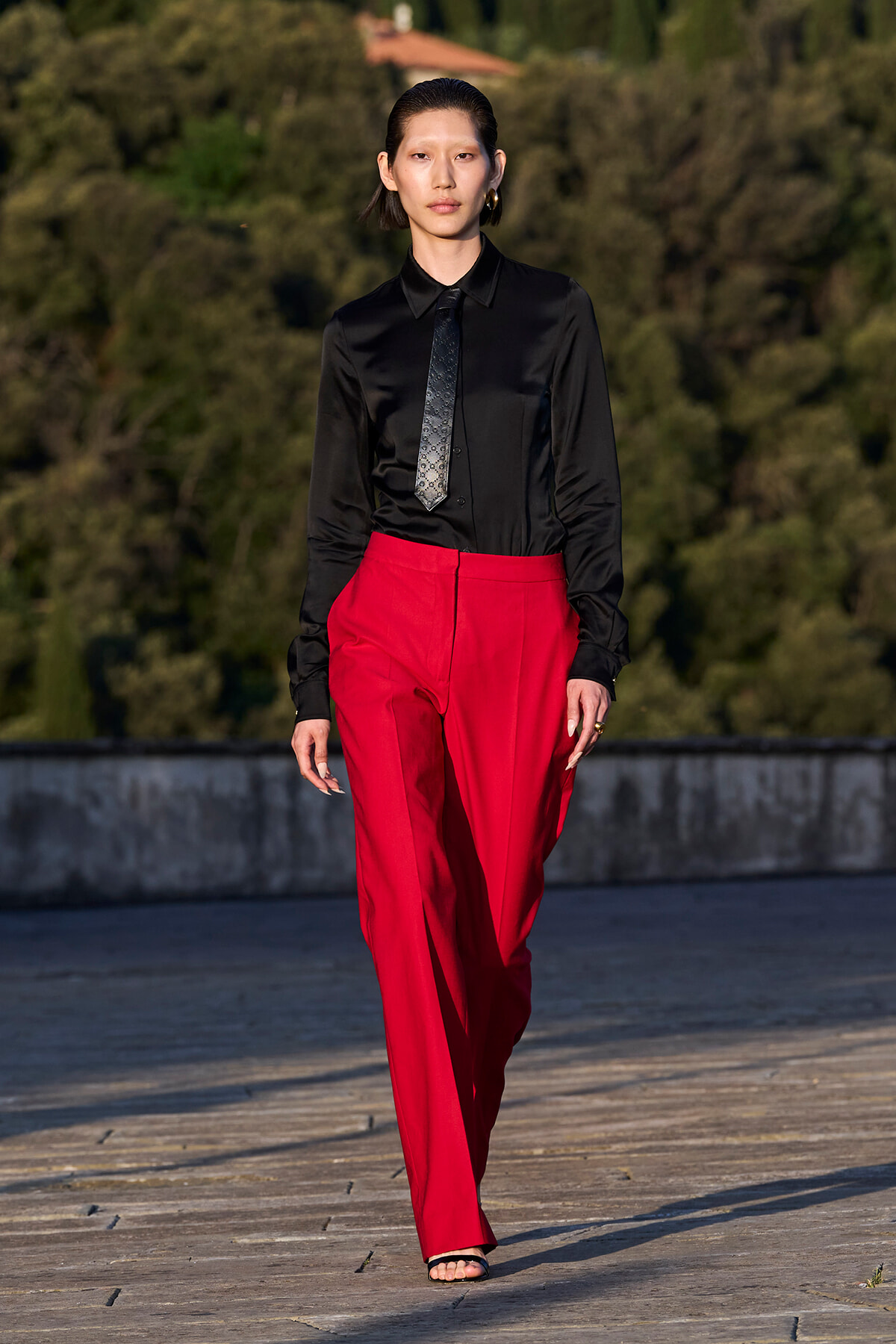 Model walks the runway in a black satin shirt, patterned tie, and bright red tailored wide-leg pants.