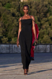 Model on an outdoor runway wearing a black halter-neck gown with a small clutch in hand.
