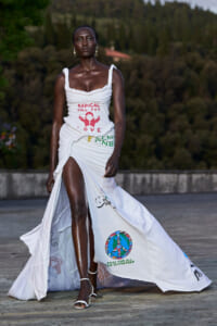 Model on a runway wearing a white ruched gown with printed activism text and a long train with logos, outdoors against a trees backdrop.