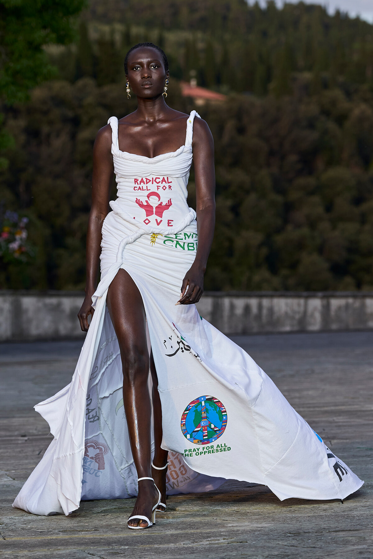 Model on a runway wearing a white ruched gown with printed activism text and a long train with logos, outdoors against a trees backdrop.