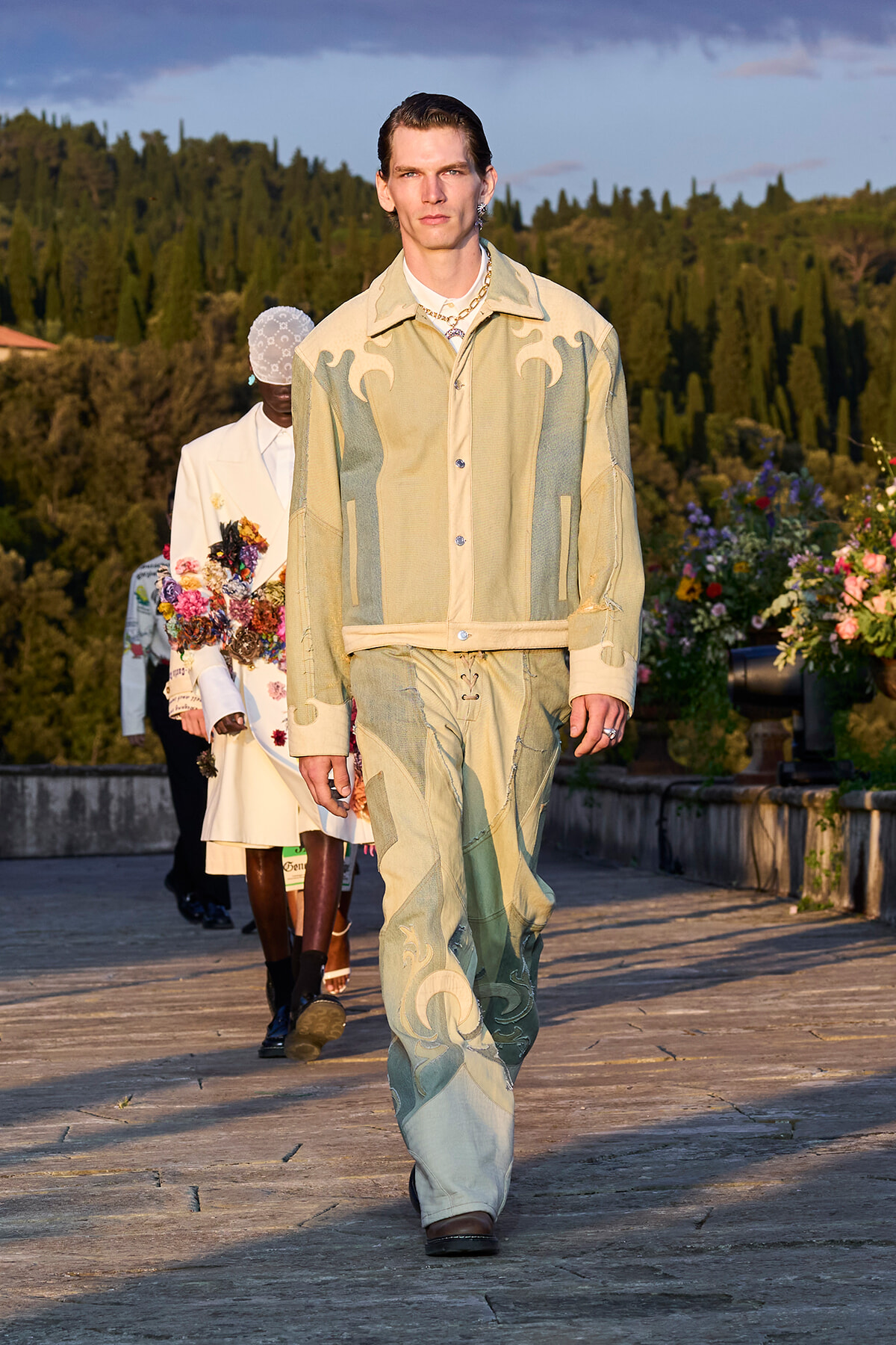 Male model walks a wooden runway outdoors in a beige-green patchwork denim outfit, with floral arrangements along the catwalk