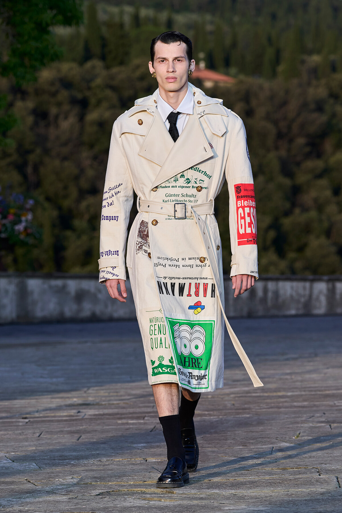 Male model wearing a beige trench coat covered with printed graphics and logos, walking outdoors on a stone terrace.