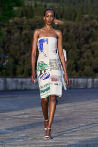 Model walking on an outdoor runway in a strapless knee-length dress covered with newspaper-style prints and graphics, paired with white ankle-strap sandals.