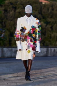 Model wearing a white double-breasted coat densely covered in multicolored fabric flowers, with a lace cap and blue shoes on a runway.