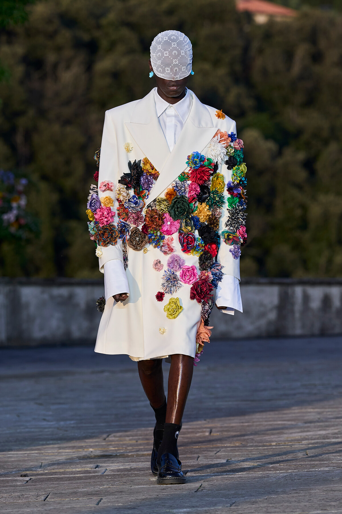 Model wearing a white double-breasted coat densely covered in multicolored fabric flowers, with a lace cap and blue shoes on a runway.