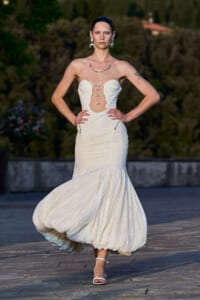 Model walking on a runway wearing a strapless white lace gown with a lace-up bodice and bubble hem, outdoors