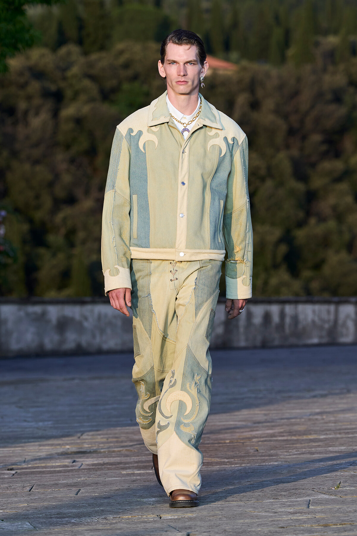 Male model walking on an outdoor runway in a pale patchwork denim suit with embroidered scrolling motifs, paired with brown boots and gold jewelry.