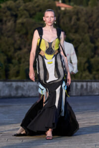 Model wearing a yellow-black survival harness over a flowing black-and-white dress, walking on stone pavement outdoors.
