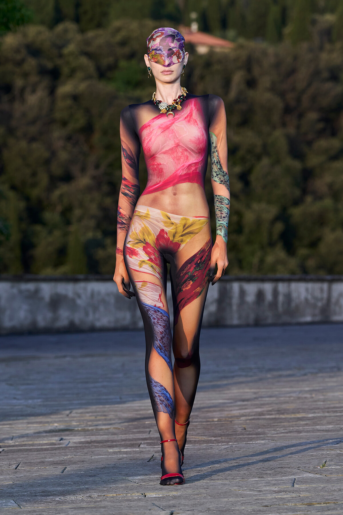 Model walking a runway in a full-body floral print sheer bodysuit with black sleeves, headscarf, and red heels.
