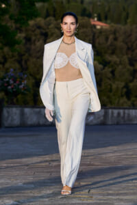 Model walking a runway in a white suit: blazer over beige lace bustier and matching trousers, outdoors with greenery in the background.