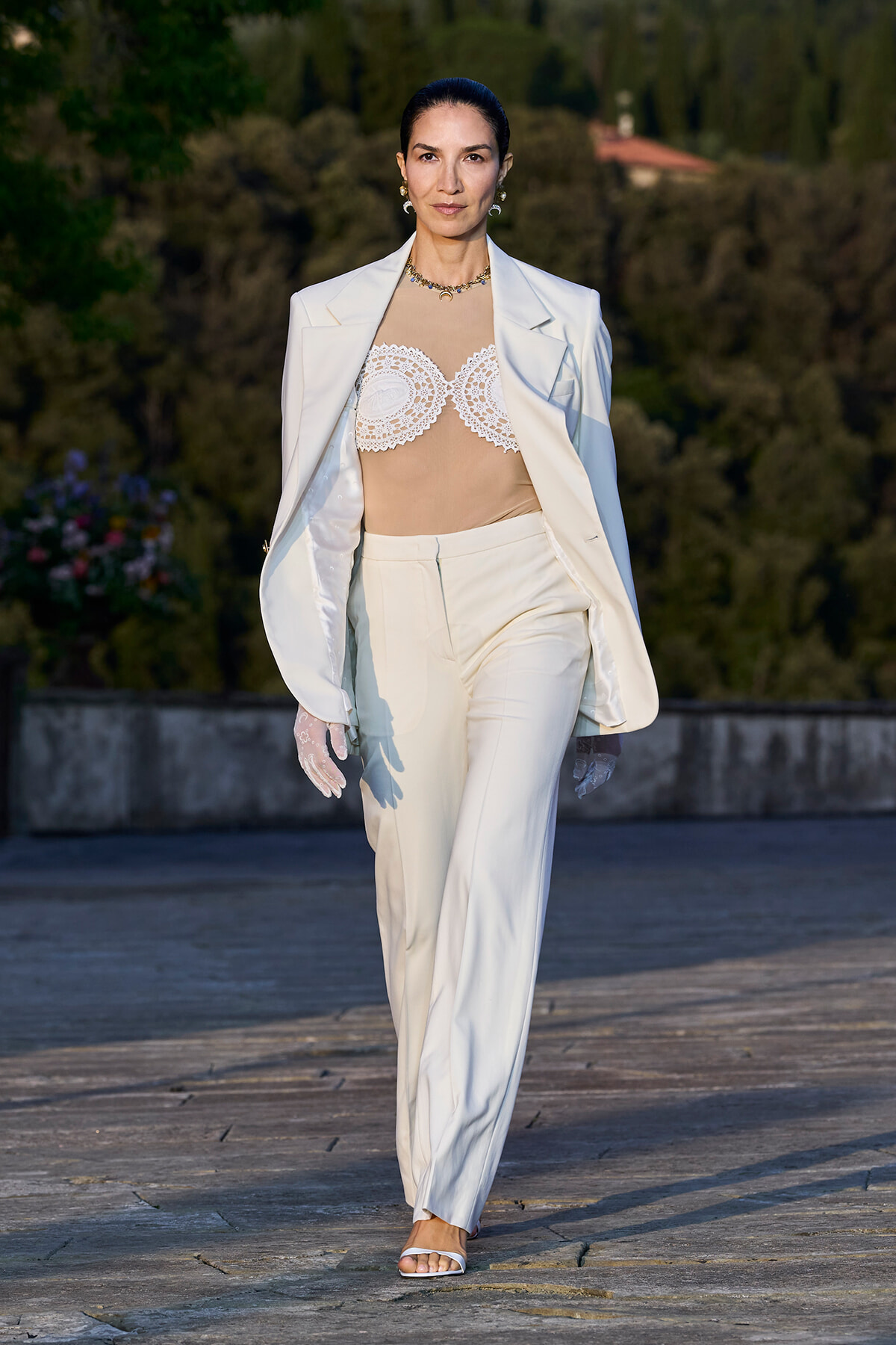 Model walking a runway in a white suit: blazer over beige lace bustier and matching trousers, outdoors with greenery in the background.