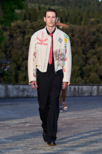 Male model walking on an outdoor runway in a cream jacket with patches, a red tie, and black pants.