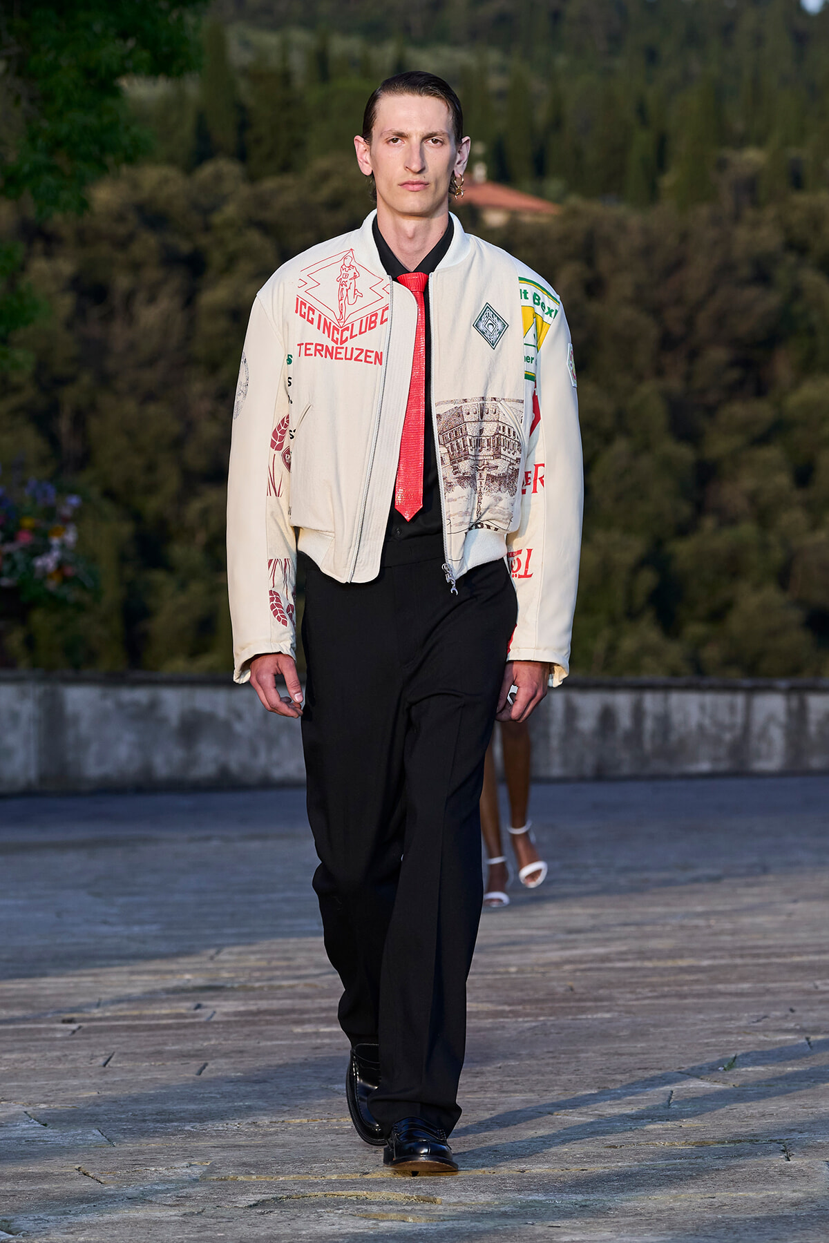 Male model walking on an outdoor runway in a cream jacket with patches, a red tie, and black pants.