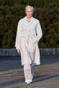 Older woman model walks an outdoor runway in a white embroidered coat with lace-trimmed pants/leggings.
