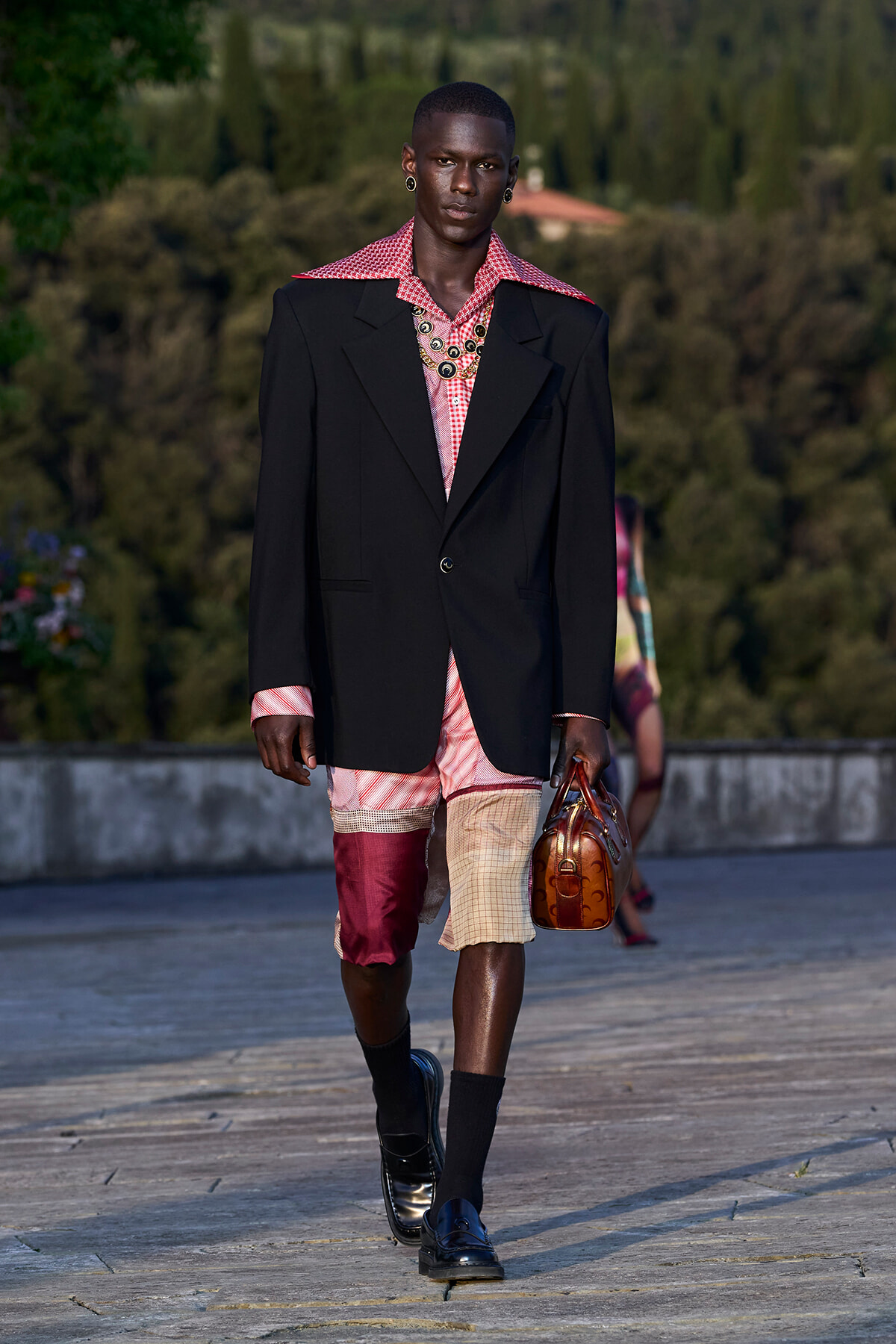 Male runway model wearing a black oversized blazer over a red patterned shirt and patchwork shorts in pink, beige, and burgundy tones, with black socks, patent loafers, and a brown leather handbag
