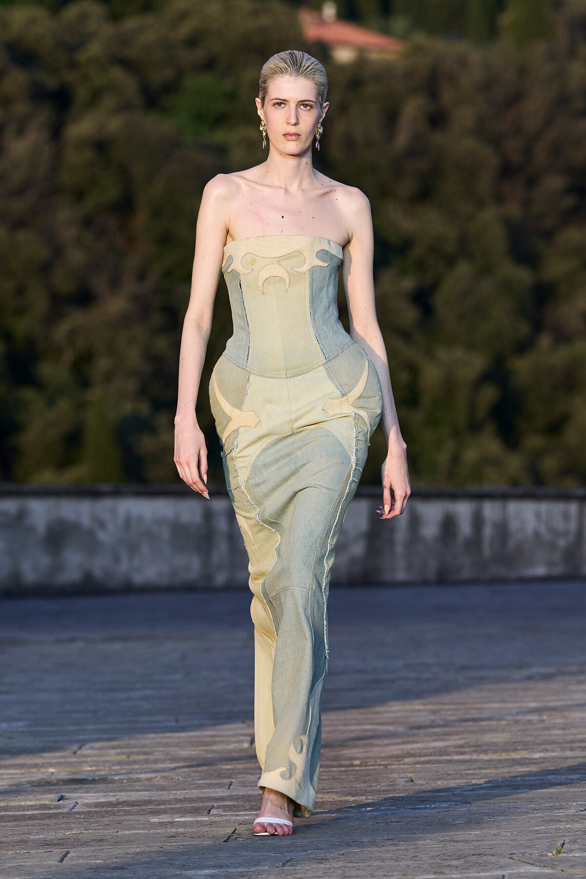 Model walking a runway in a strapless, patchwork denim gown with pale yellow panels, outdoors on a stone catwalk.