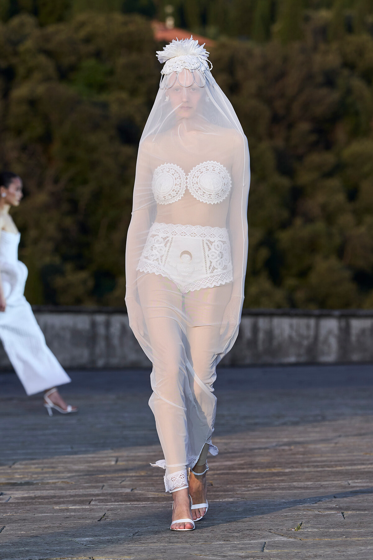 Model walking a runway in sheer bridal ensemble: lace pasties, high-waisted lace shorts, veil, and headpiece, white strappy heels; outdoor fashion show setting