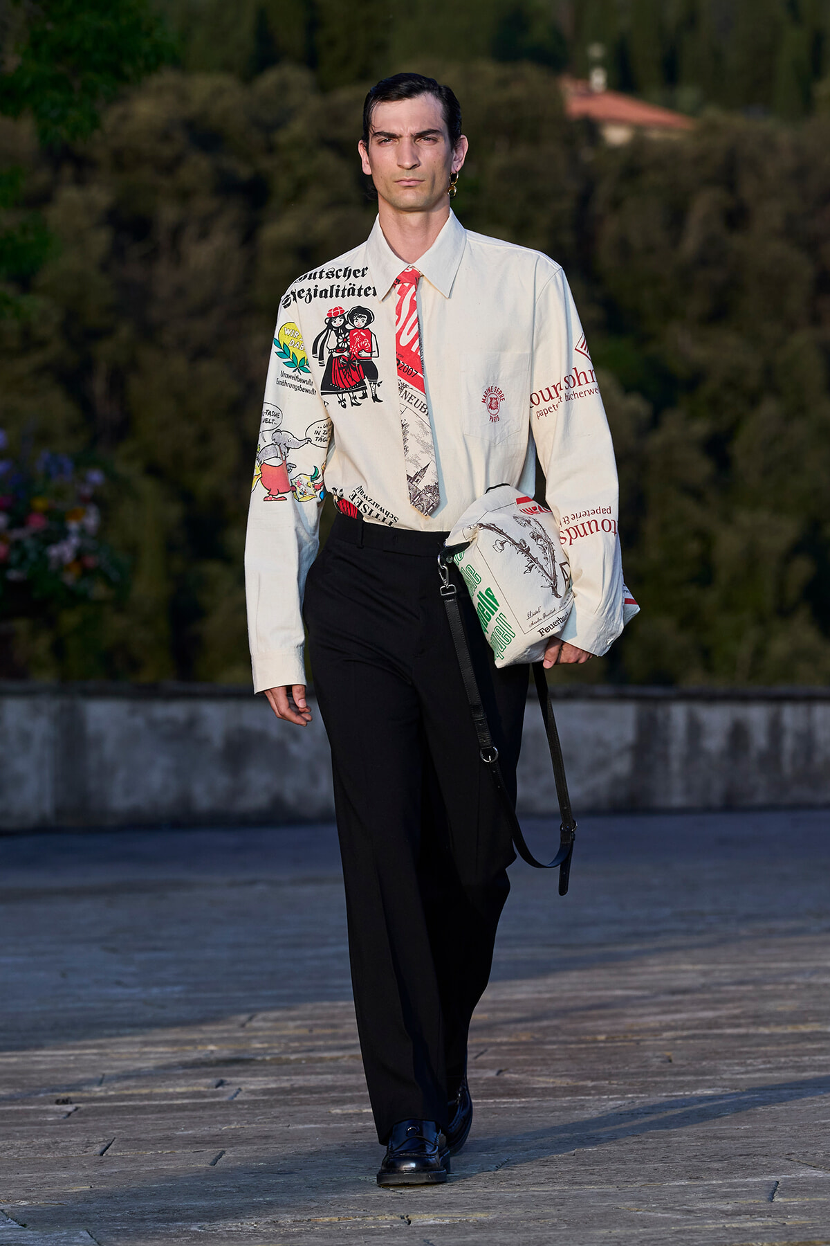 Male model walks the runway in a white graphic shirt, black trousers, blue shoes, carrying a white bag with a strap under his arm.