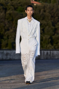 Male model in an all-white suit with lace embroidery walks on an outdoor runway, gold necklace adding contrast.