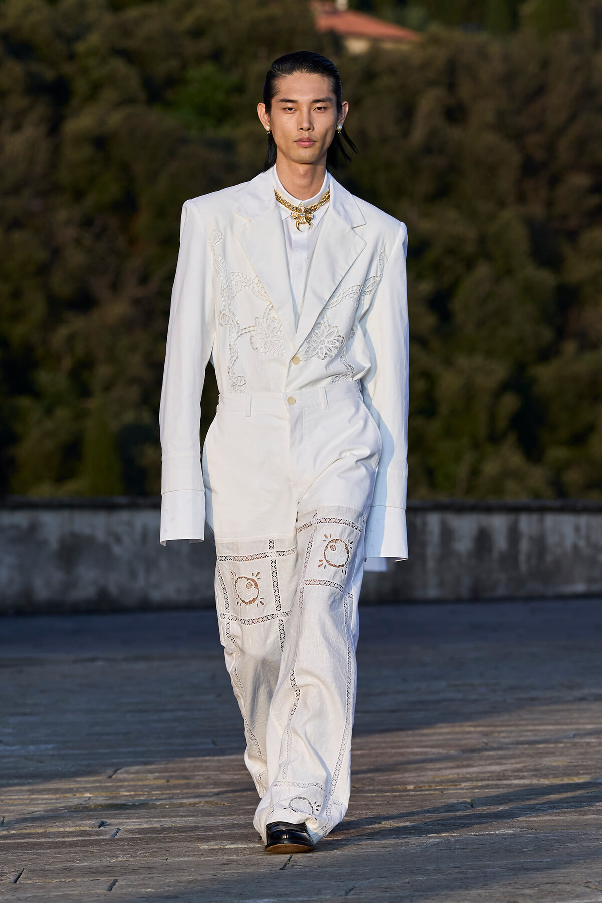 Male model in an all-white suit with lace embroidery walks on an outdoor runway, gold necklace adding contrast.