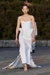 Model walks the runway in a strapless white couture gown with a textured ribbed pattern and sculpted side hips, trailing a long train.