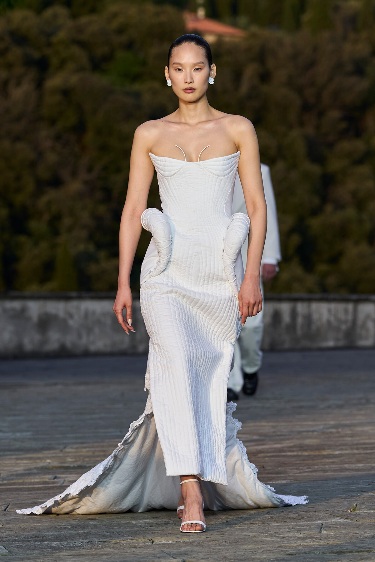 Model walks the runway in a strapless white couture gown with a textured ribbed pattern and sculpted side hips, trailing a long train.