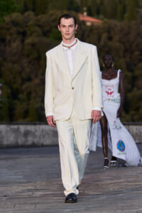 Male model walks a runway in a cream-white oversized suit with a white shirt and red beaded necklace at an outdoor fashion show.