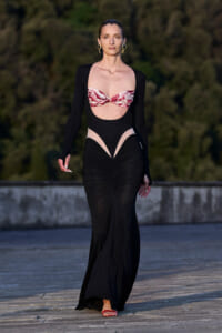 Model walking a runway in a black cutout gown with a pink patterned bandeau top and gold hoop earrings.