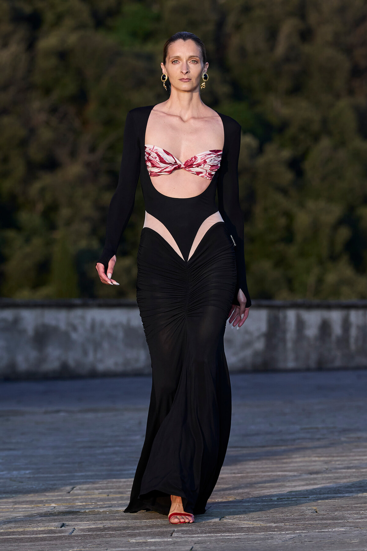 Model walking a runway in a black cutout gown with a pink patterned bandeau top and gold hoop earrings.