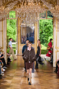 Male model walks the runway in a brown leather jacket and burgundy shorts inside a gilded, chandelier-lit room with spectators on both sides.