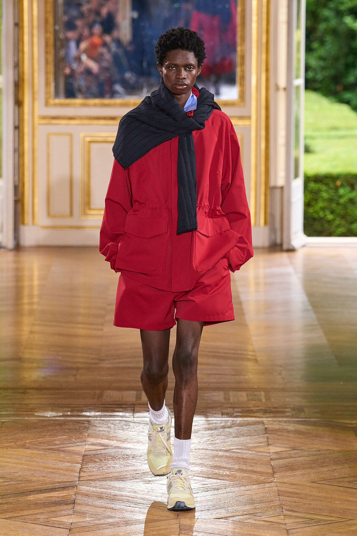 Model walking a fashion runway in a red two-piece set with a black scarf draped over shoulders inside a gilded, ornate room.